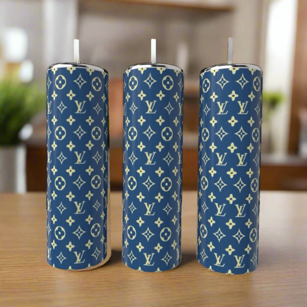Three cylindrical candles with blue exteriors and white Louis Vuitton monogram patterns stand upright on a wooden surface, paired with a Kreative Kreationz LV Blue & Yellow 20oz Tumbler in the softly blurred indoor background.