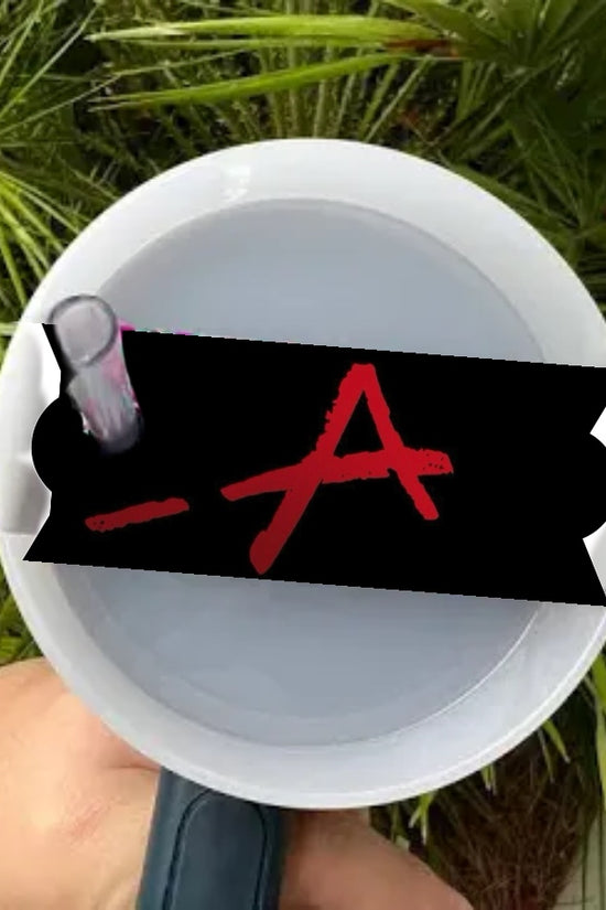 A person holds a white megaphone with a black sticker displaying a red, hand-drawn 'A', alluding to Pretty Little Liars. Behind it, green palm leaves enhance the intrigue. The sticker is the 