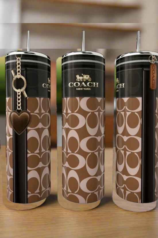 Three Coach Buckle 20oz Skinny Tumblers by Kreative Kreationz, featuring brown/beige logos, black stripe, metal lids, and straws, sit on a wooden surface; one stainless steel tumbler comes with a heart-shaped keychain.