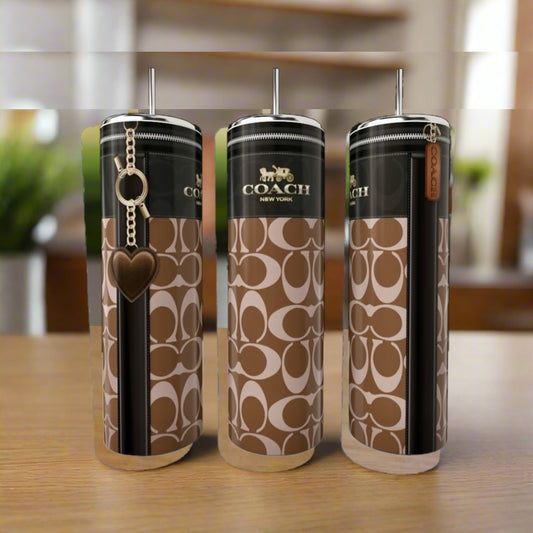 Three Coach Buckle 20oz Skinny Tumblers by Kreative Kreationz, featuring brown/beige logos, black stripe, metal lids, and straws, sit on a wooden surface; one stainless steel tumbler comes with a heart-shaped keychain.