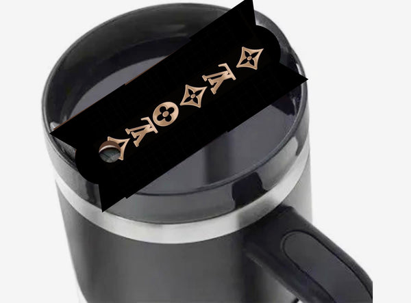 A black Stanley tumbler is topped with the Kreative Kreationz Louis Vuitton | Stanley Lid Plate/Topper, a designer lid featuring gold-colored LV symbols that partially covers the mug’s opening.