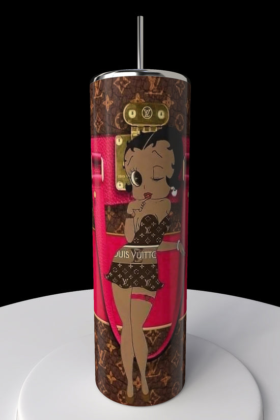 The Betty B Coach 20oz Skinny Tumbler by Kreative Kreationz showcases a cartoon character in a stylish outfit on a designer-themed brown and pink background. It comes with a metal straw and is elegantly displayed on a white surface against a black backdrop.