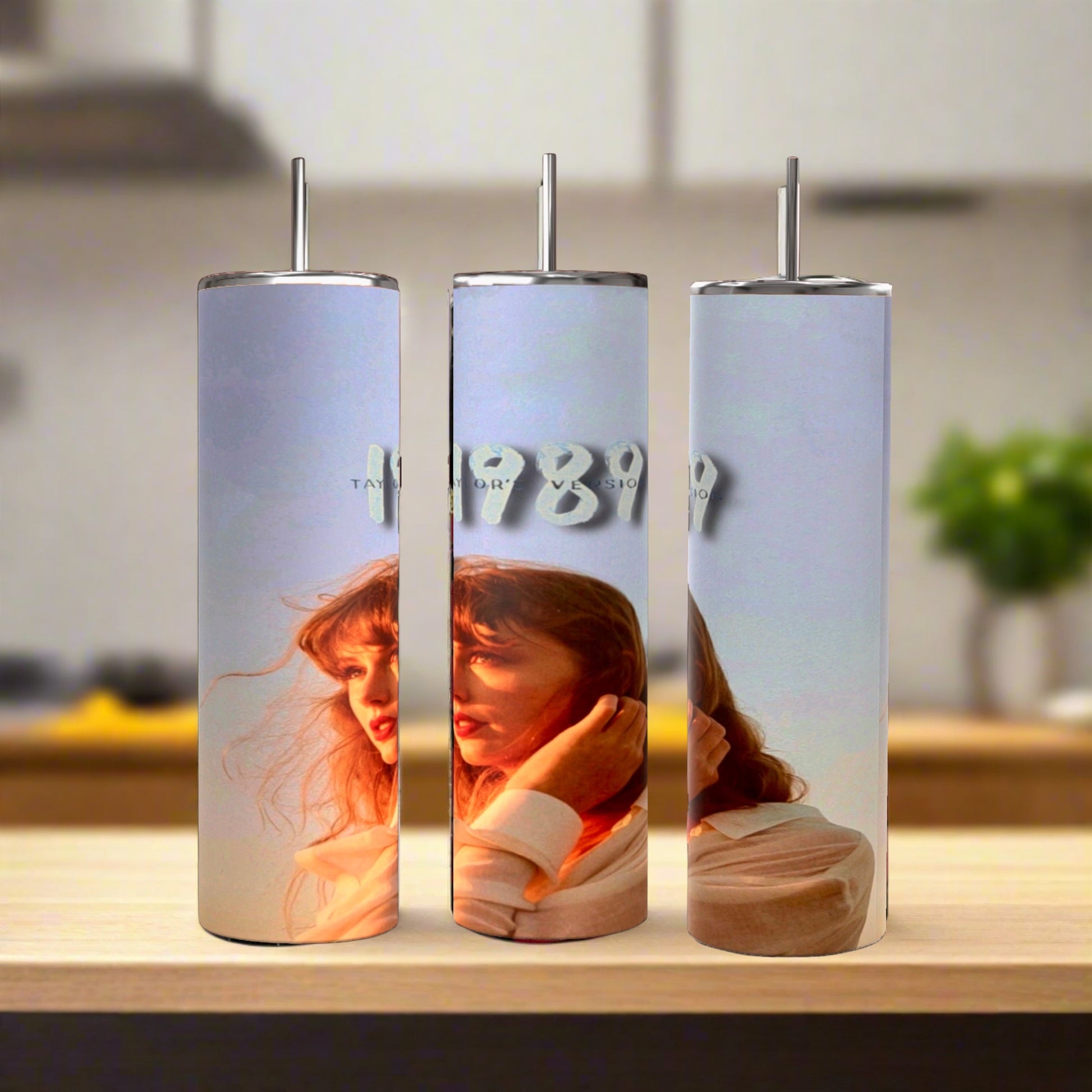 Three Kreative Kreationz Taylors Version 1989 20oz Tumblers with straws are displayed on wood. Each eco-friendly stainless steel tumbler shows a woman with red hair in sunlight, and "1989" in silver text appears on the center one. Blurred kitchen background.