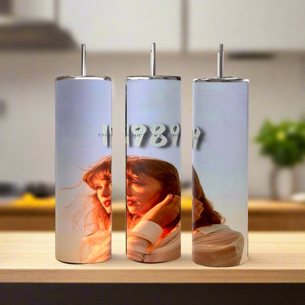 Three Kreative Kreationz Taylors Version 1989 20oz Tumblers with straws are displayed on wood. Each eco-friendly stainless steel tumbler shows a woman with red hair in sunlight, and "1989" in silver text appears on the center one. Blurred kitchen background.