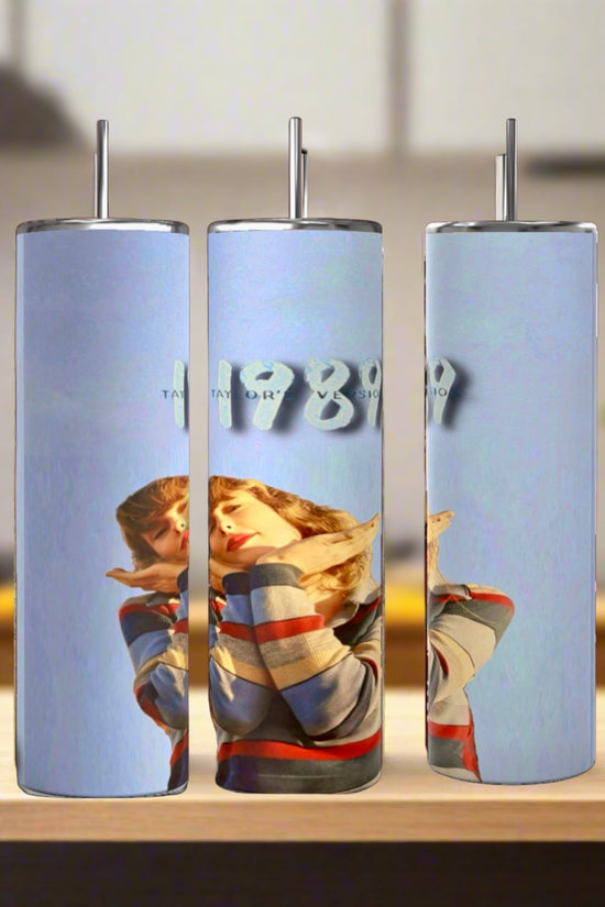 Three Kreative Kreationz Taylor Swift 1989 20oz stainless steel tumblers with metal straws sit on a wooden surface. Each eco-friendly, reusable tumbler shows a woman in a colorful sweater and “1989” on a light blue background.