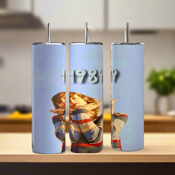 Three Kreative Kreationz Taylor Swift 1989 20oz stainless steel tumblers with metal straws sit on a wooden surface. Each eco-friendly, reusable tumbler shows a woman in a colorful sweater and “1989” on a light blue background.