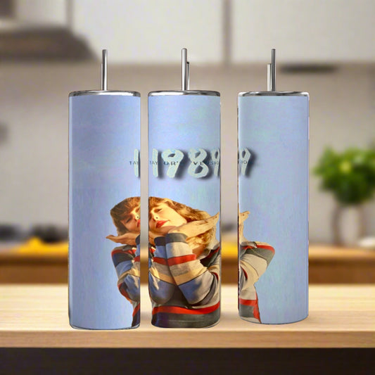 Three Kreative Kreationz Taylor Swift 1989 20oz stainless steel tumblers with metal straws sit on a wooden surface. Each eco-friendly, reusable tumbler shows a woman in a colorful sweater and “1989” on a light blue background.