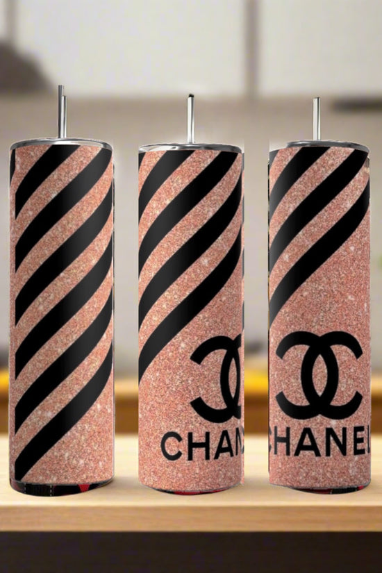 Three Chanel Pink Stripe 20oz Skinny Tumblers by Kreative Kreationz, each with a metal straw, are displayed on wood. The insulated tumblers feature rose gold glitter, black stripes, and the CHANEL logo. A softly blurred kitchen is in the background.