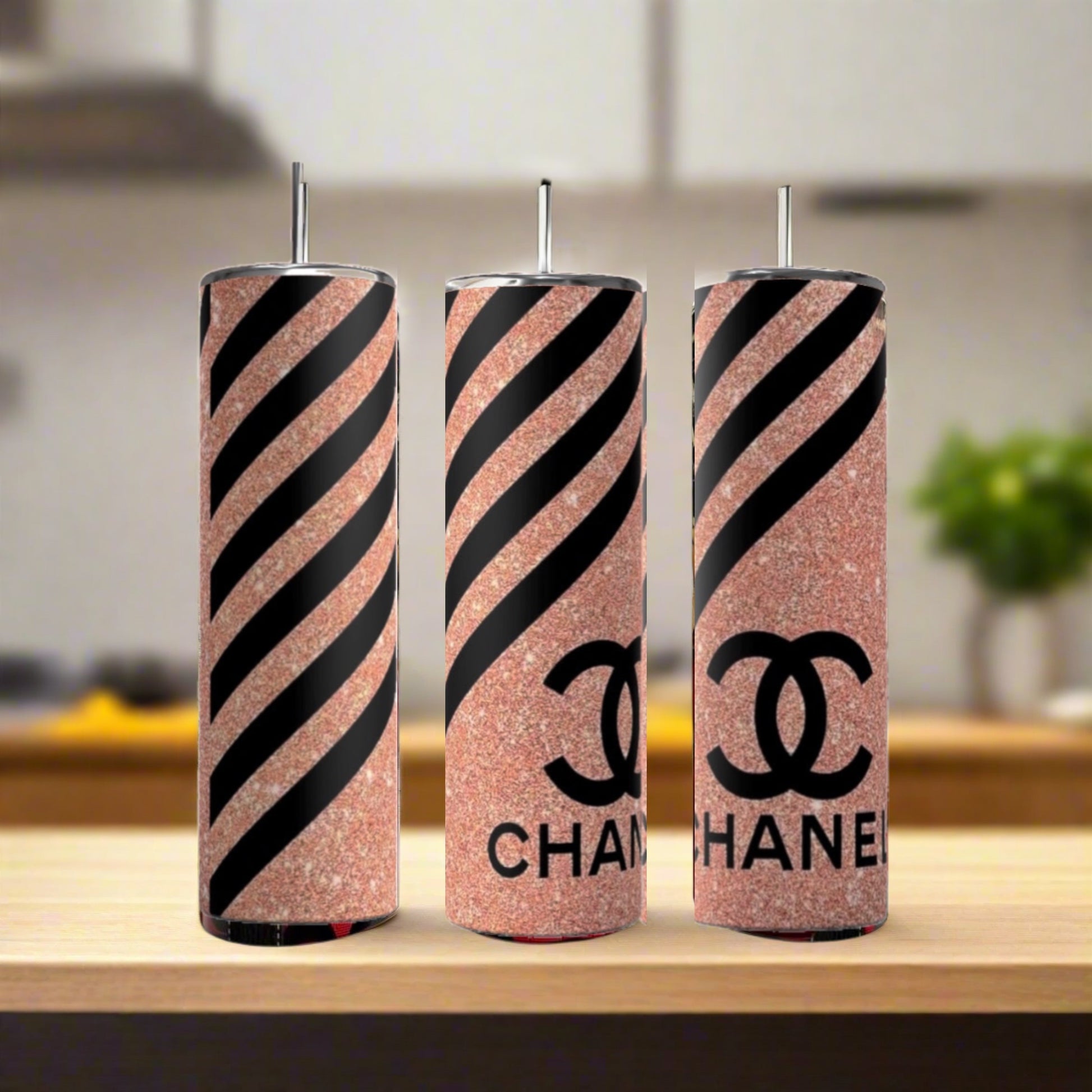 Three Chanel Pink Stripe 20oz Skinny Tumblers by Kreative Kreationz, each with a metal straw, are displayed on wood. The insulated tumblers feature rose gold glitter, black stripes, and the CHANEL logo. A softly blurred kitchen is in the background.