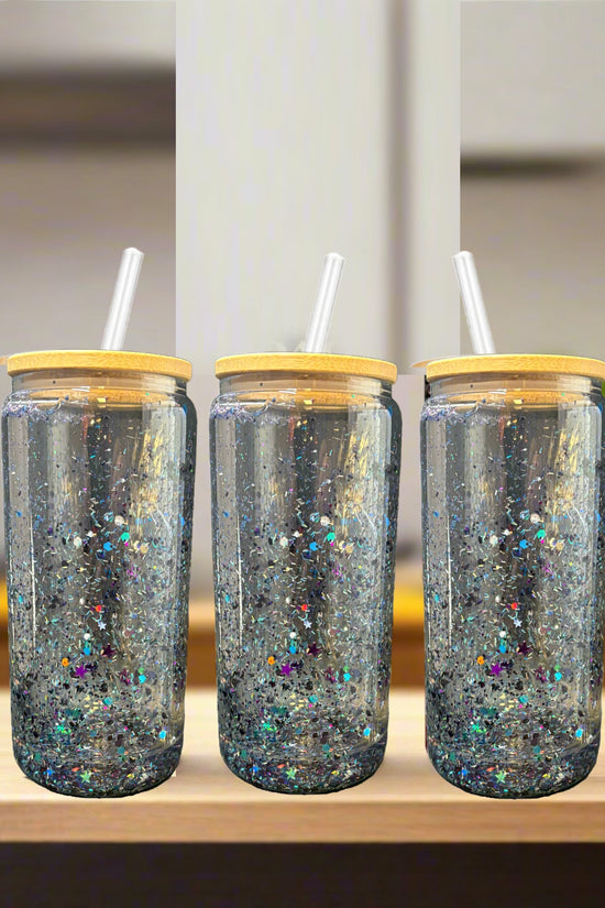 Three Kreative Kreationz Snowglobe Tumblers—16oz clear glass with bamboo lids and straws—are filled with sparkling glitter and sit side by side on a light wooden surface, against a blurred kitchen background.