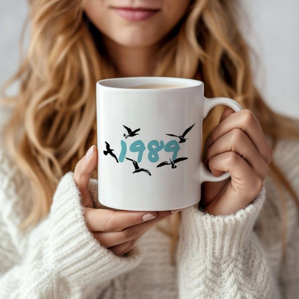 A woman with long, wavy blonde hair holds a Kreative Kreationz 1989 Ceramic Mug, featuring "1989" in blue-green text and flying black birds. She wears a cozy white sweater—a perfect Swiftie gift.